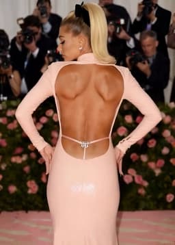The woman stands with her back to the viewer, hands placed on her hips and elbows angled outward, her posture drawing the light pink fabric of the long-sleeved dress taut across her shoulders and waist. The dress features a high neckline in front that continues into a dramatic open back, the fabric cut away in a wide, curving shape that exposes the full expanse of her tanned skin from shoulder blades to lower back. A single thin pink strap spans the open section just above the waist, secured at the center by a sparkling jeweled clasp that sits directly against her spine, with a thin white string running straight down from beneath it and slipping between the upper curves of the buttocks. The dress clings closely to her form, its smooth, slightly glossy texture outlining the natural contours of her back and the gentle inward taper at the waist before flaring smoothly over the hips. Subtle sparkles dot the lower portion of the skirt visible at the base of the frame. Her blonde hair is pulled into a high, sleek ponytail secured by a large black bow, with the tail cascading down one side. Small pearl stud earrings catch the light at her ears, and her hands rest with fingers lightly curled against the dress fabric. The open back design frames the exposed skin, accentuating its smooth surface and the subtle lines of muscle beneath.