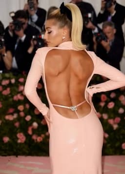The woman stands with her back to the viewer, head turned in profile to reveal a sharp jawline and dark eyeliner along her lashes. Her blonde hair is pulled into a high, sleek ponytail secured by a large black bow, with the tail cascading down one shoulder. The pale pink dress clings closely to her frame, its glossy fabric catching light along the curve of her hips and the small of her back. A wide oval-shaped opening exposes nearly the entire back, framed by narrow sleeves that extend to the wrists and a high collar visible at the nape. Smooth skin stretches over defined muscle contours and subtle ridges along the spine, with a faint sheen catching light across the planes of her shoulder blades and the gentle inward curve at the waist. A thin pink band stretches horizontally across the lower back, secured at center with a rectangular jeweled clasp whose facets glint against the skin. Her posture places both hands on her hips, elbows angled outward, which draws the open edges of the dress slightly apart and accentuates the contoured planes of her back and the gentle taper at the waist. A small pearl stud earring is visible in the ear turned toward the viewer. The fitted skirt portion molds tightly over the hips, its sheen highlighting subtle surface reflections without additional adornment.