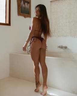 She stands with her back toward the viewer, weight shifted onto one leg so her hips tilt and the rounded curve of her backside is pronounced, while she glances over her shoulder with a bright smile. Her long, sun-warmed hair falls down her back in loose waves. A deep burgundy string bikini covers her, the top tied across her upper back and the bottoms secured by thin ties that knot at each hip and trail downward. Her thighs are full and powerfully shaped, the skin smooth and evenly tanned, tapering to noticeably slender ankles adorned by a delicate gold chain on the right. Both feet are completely bare, toes pressing lightly against the floor; the left foot reveals neatly painted toenails in a vivid blue gloss. A small dark tattoo sits just above her left ankle bone, and another appears higher on the outer thigh. Thin gold bracelets rest on her left wrist, and her arms hang relaxed at her sides. The overall stance presents a strong, feminine silhouette with clear emphasis on the toned length of her legs and the exposed, polished detail of her feet.