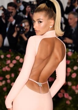 The woman stands with her back toward the viewer, her torso twisted slightly so that her face appears in profile over one shoulder. She wears a form-fitting pale pink gown with long sleeves and a high, close neckline that contrasts with the dramatic open back. A large cutout extends from just below the shoulder blades down to the base of the spine, exposing smooth, evenly tanned skin and the defined contours of her upper and mid-back muscles. A single narrow pink strap crosses the lower back, fastened at the center with a small jeweled ornament whose facets catch the light. Directly below, a thin pink thong string runs downward between the rounded cheeks before disappearing into the dress opening. The fabric clings closely to the curve of her hips and the rounded shape of her buttocks, creating a continuous, unbroken line from waist to hem. Her blonde hair is pulled into a high, sleek ponytail secured by a wide black bow, the tail falling forward over one shoulder. Her posture, with hands resting at her sides and elbows angled outward, draws attention to the narrowness of her waist relative to the flare of her hips and the expanse of bare back framed by the dress.