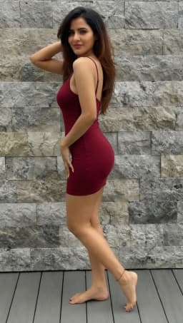 The woman wears a tight maroon dress that clings closely to her torso and hips, its thin straps resting on her shoulders and the hem ending high on her thighs. She stands with her body angled away, weight shifted onto one leg while the other extends slightly forward, one hand lifted to touch her long, brown hair and the other resting near her hip. Her legs appear firm and rounded, the skin smooth and evenly toned, leading down to fully bare feet. A thin black corded anklet encircles her left ankle, and both sets of toenails are coated in a glossy, black polish that contrasts with the natural color of her skin. Her feet are positioned arched, raised high with toes bearing her weight, her arches visible, the bare, wrinkled soles fully exposed. The dress fabric stretches across her lower back and hips, outlining their curves in a continuous line from waist to thigh.