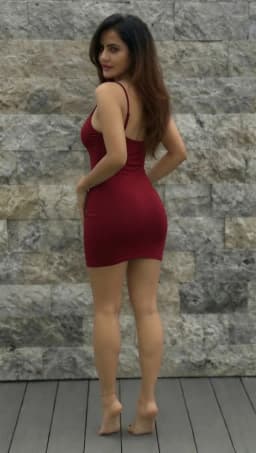 The woman stands with her back angled toward the viewer, head turned to glance over her left shoulder, dark hair falling in loose waves across her upper back. Her form-fitting red dress clings closely to her torso and hips, supported by thin straps that cross her shoulders and leave much of her back exposed. Her hands rest lightly on her upper thighs, accentuating the curve of her waist and the rounded contour of her hips. Her bare legs extend downward with smooth, even skin tone and a defined feminine muscularity along the calves and thighs, leading to slender ankles. A delicate black cord anklet wraps around her left ankle. Both feet are completely bare, their arches and toes visible against the floor, the skin appearing soft and light against her sensual, glossy, black toenail polish. The posture highlights the dress’s hem riding high on her thighs and the natural alignment of her stance, drawing attention to the length and proportion of her legs.