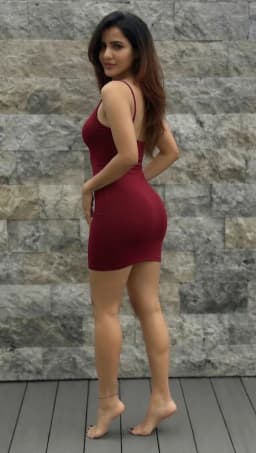 The woman stands with her weight shifted onto one leg, hips angled away while she glances back over her shoulder, one hand resting on her hip. She wears a tight-fitting maroon mini dress with narrow straps that follow the lines of her shoulders and frame the open back, the fabric stretched smoothly over her bust, waist, and hips before ending high on her thighs. Her bare legs appear full and toned, the skin smooth and evenly tanned, with the short hem leaving the full length from mid-thigh downward exposed. A thin black, corded anklet rests around her left ankle. Both feet are completely bare, their naked soles and arches visible as they rise arched, toes painted in a glossy, black polish that matches the depth of the dress. A small tattoo shows on the inner forearm near the wrist.