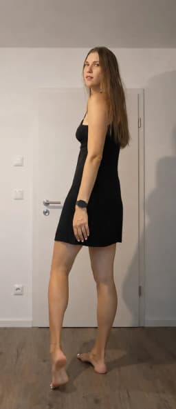 A woman stands barefoot, and wears a short black sleeveless dress with thin straps. Her body faces away from the camera at a three-quarter angle, while her head is turned over her left shoulder to face the viewer. Long straight light-brown hair falls past her shoulders. Her left arm hangs at her side with the hand resting near her thigh; a black fitness tracker is on her left wrist and white nail polish is visible on her fingernails. A thin gold anklet is on her right ankle, and sexy, bold, white nail polish appears on her toes. Her right foot is flat on the floor; her left foot is raised with the heel lifted and toes pointing downward.