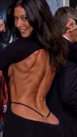 The woman stands with her back angled toward the viewer, her torso twisted so she can look over her left shoulder. Her face is turned toward the camera, mouth open in a smile that shows her upper teeth, eyes bright and directed forward. Long, dark brown hair falls in loose waves across her right shoulder and down her back, partially covering the right side of her exposed skin.

She wears a black garment with long sleeves that leaves the entire back open from the neck down. Thin black straps of her visible thong cross the lower back, adorned in their center by a small gold ring embellishment where the thong connects downward between her buttocks cheeks beneath the garment, while a fine gold chain hangs vertically from the upper back, ending in a small pendant dangling precipitously in front of the sensual curve of her lower back. The fabric of her dress clings closely at the shoulders and sides, revealing the full width of her back and the way the skin follows the underlying shape with clear ridges and separations across the upper and middle sections.

Her left arm is raised and bent, with the hand partially visible near the side of her head, showing a small ring and manicured nails. The right side of her back remains fully visible, the skin smooth and evenly toned under the open cut of the garment.
