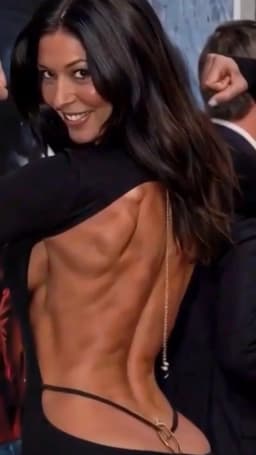 She turns her upper body toward the viewer while looking back over one shoulder, her face lit by a broad smile that shows her teeth. Long, dark hair falls in loose waves across her back and one shoulder. The black garment she wears is cut low and open down the center of her back, held in place by a slender gold chain that drapes vertically along one side. At the lower curve of her back, the horizontally reaching narrow black straps of her thong are visible at either side, encircling the base of her back and adorned with a small gold ring embellishment where the strap passes beneath the bottom hem of the garment to continue between her cheeks. The exposed skin of her back and sides displays smooth, contoured shapes and firm lines that follow the natural curves of her torso.