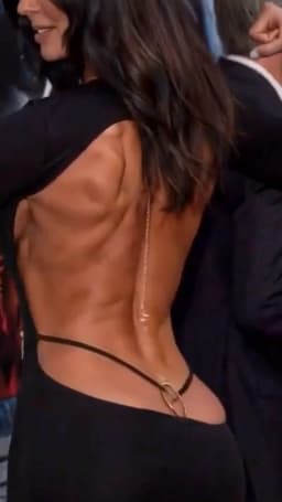 The woman stands with her back turned toward the viewer, her head angled slightly to one side so her profile appears, lips parted in a gentle smile. Long, dark wavy hair tumbles down her back and over one shoulder, partially covering the upper portion of the open dress. The black garment fits closely to her frame, its back cut away in a wide, deep opening that leaves most of the skin exposed from shoulders to lower waist. A slender gold chain runs straight down the center of the exposed area, while the horizontally-reaching narrow black straps of her thong are visible at either of her sides, encircling the base of the back and adorned with with a small gold ring embellishment where the strap of the thong goes beneath the bottom hem of her garment to split between her butt cheeks underneath. The skin across the open expanse shows clear, sculpted contours and smooth transitions between raised and recessed planes, giving the back a distinctly defined shape.