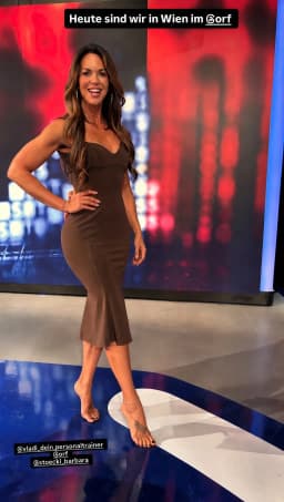 A woman stands centered in the frame on a glossy blue studio floor with white and gray geometric patterns. She wears a form-fitting brown sleeveless minidress with a sweetheart neckline and a flared hem below the knees. Her left hand rests on her hip with fingers visible; her right arm hangs at her side. She is barefoot, with glossy and seductive, black nail polish on her toes. A thin gold chain anklet is on her right ankle, and a small dark tattoo appears on the top of her right foot. Her long wavy brown hair with lighter highlights falls past her shoulders. She faces slightly toward the camera with an open-mouthed smile.